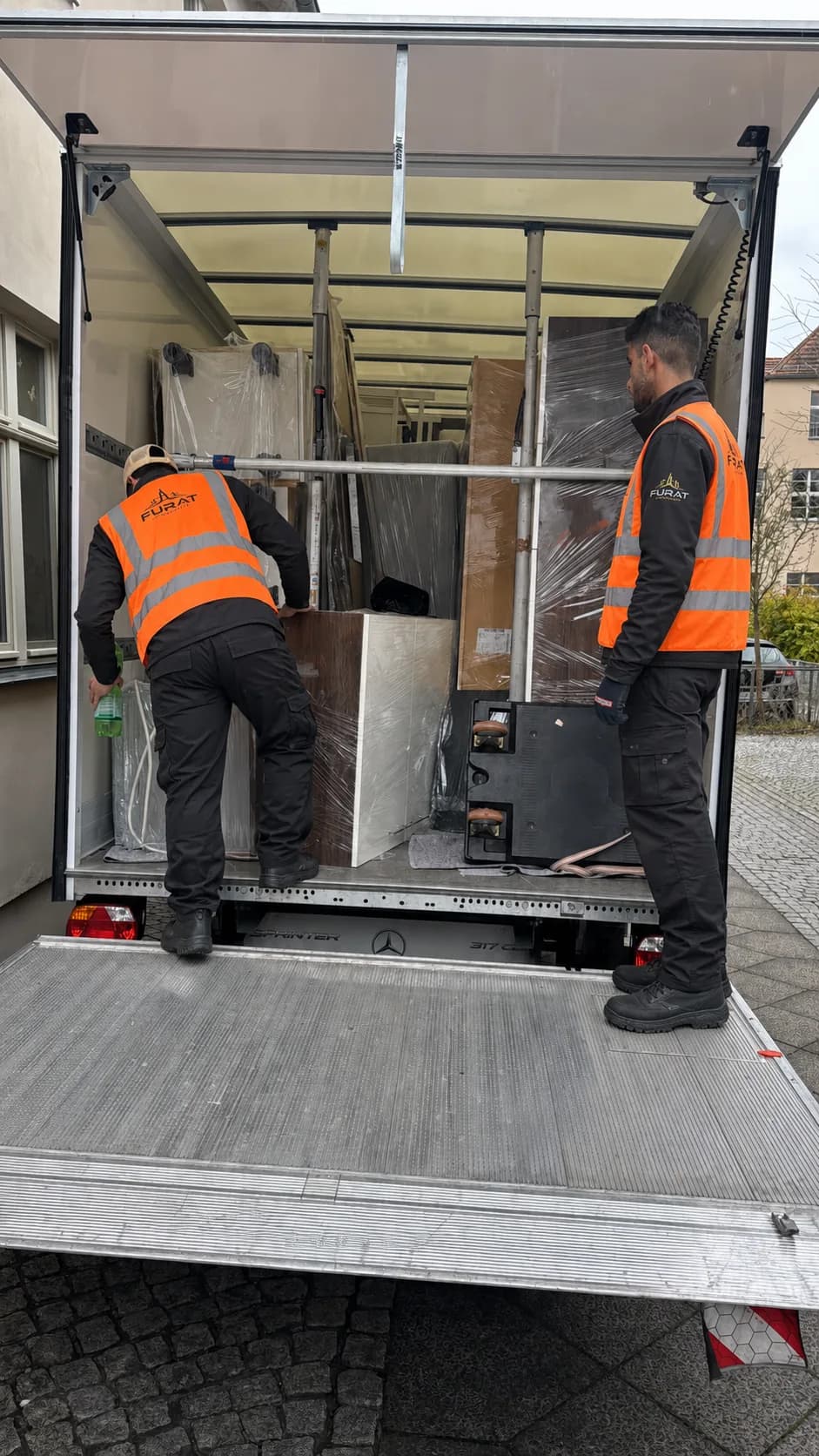 Zwei Umzugshelfer in orangefarbenen Furat-Warnwesten verstauen verpackte Möbel im Koffer-Lkw