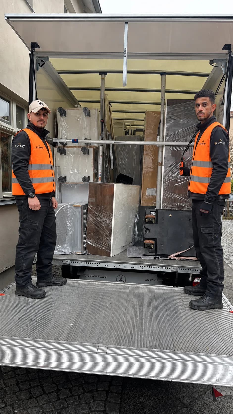Zwei Umzugshelfer in Furat-Warnwesten auf der Ladebordwand vor dem beladenen Koffer-Lkw